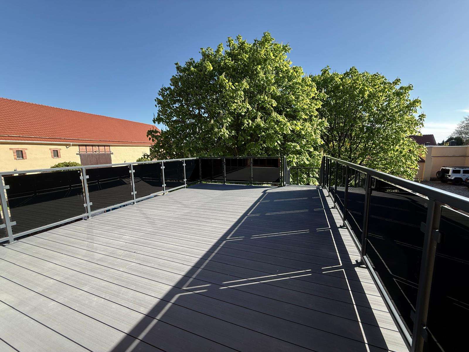 Rooftop terrace with metal-and-glass railing, gray wooden decking, and large green trees beyond under a clear blue sky.