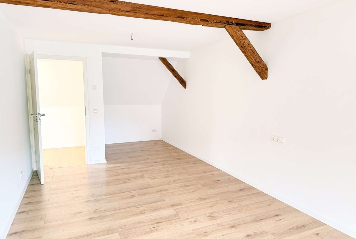 Bright, empty room with white walls, exposed rustic ceiling beams, and light wood flooring.