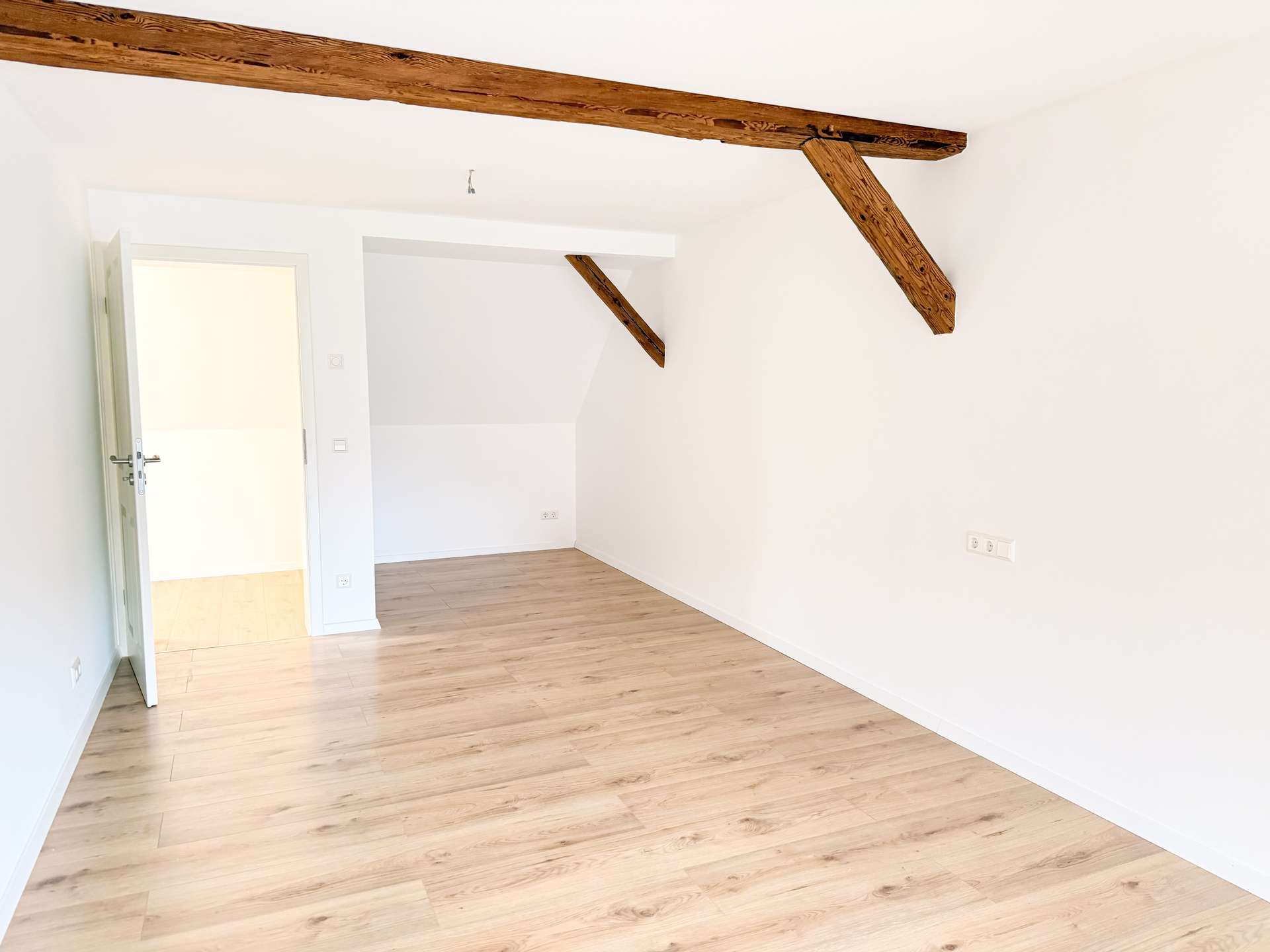 Bright, empty room with white walls, exposed rustic ceiling beams, and light wood flooring.
