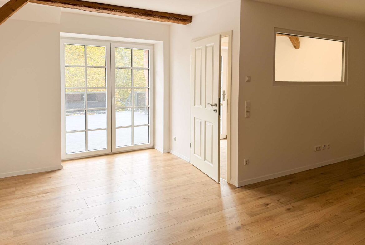 Bright, empty living space with white walls, light wood floors, and tall French doors opening to an outdoor area.