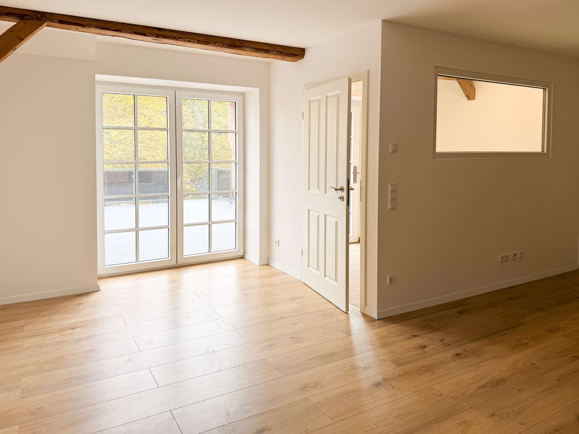 Bright, empty living space with white walls, light wood floors, and tall French doors opening to an outdoor area.