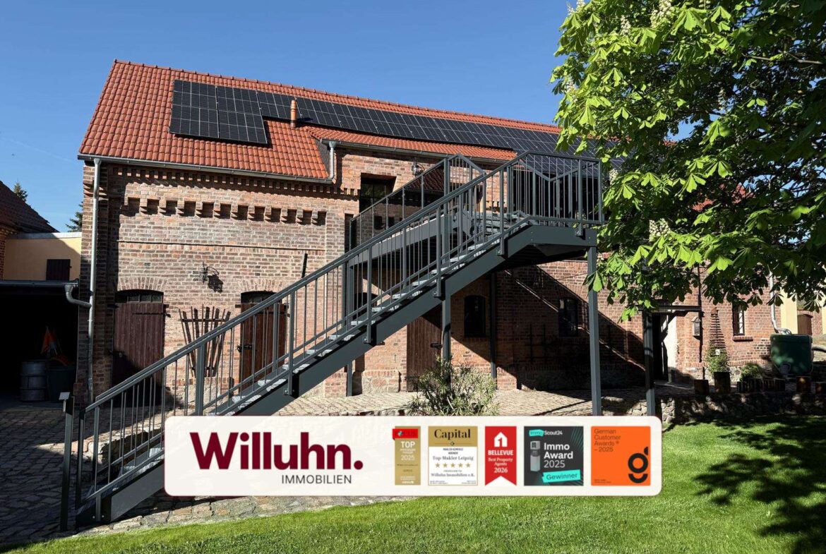 Brick building with red tile roof and solar panels, a metal exterior staircase, and a Willuhn Immobilien sign with awards in the foreground on a sunny day.
