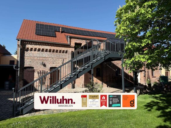 Brick building with red tile roof and solar panels, a metal exterior staircase, and a Willuhn Immobilien sign with awards in the foreground on a sunny day.