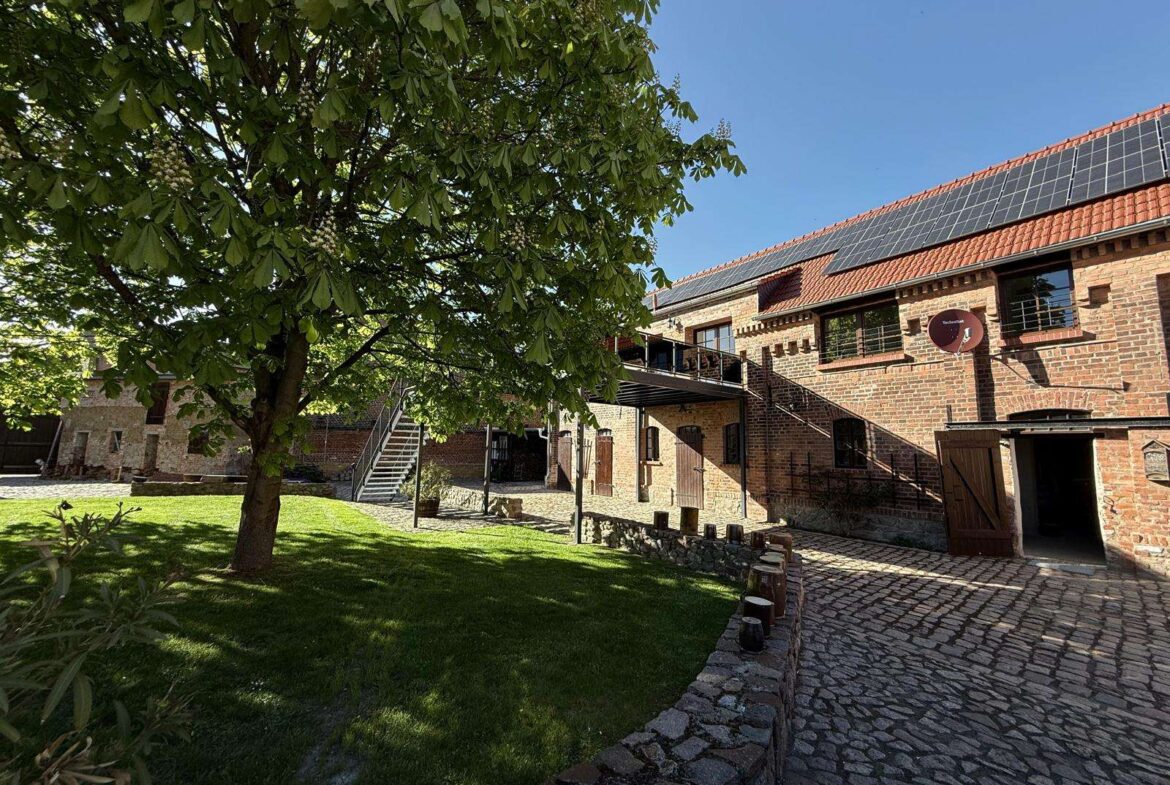 Brick building with solar panels, courtyard, and a large leafy tree casting shade on a cobblestone path.