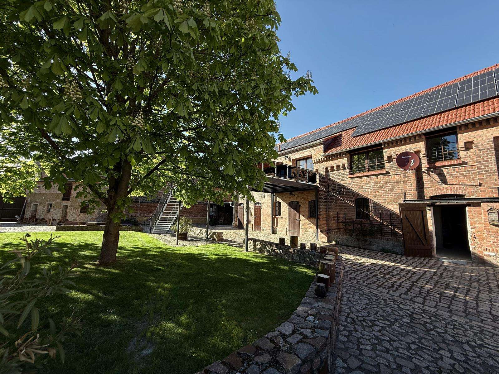 Brick building with solar panels, courtyard, and a large leafy tree casting shade on a cobblestone path.