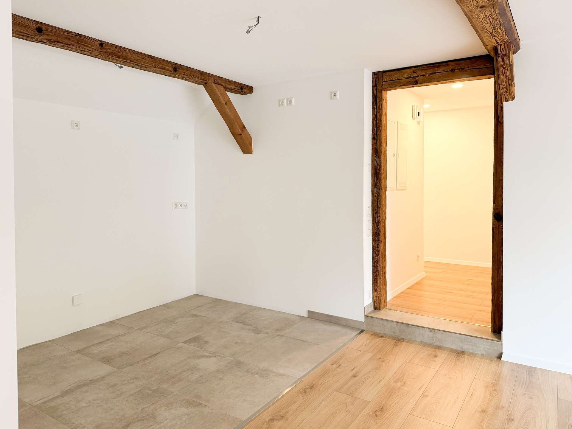 Room with white walls, exposed timber ceiling beams, and a dark wood doorway frame into a sunlit hallway.