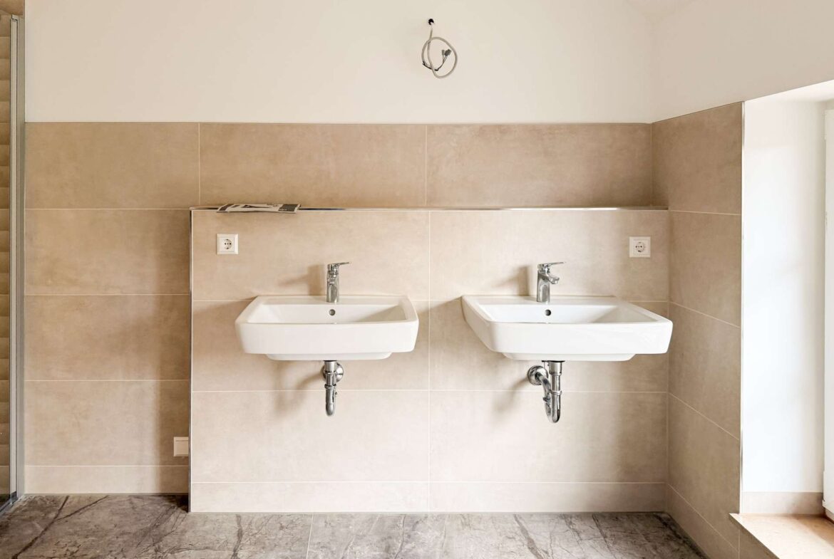 Two white wall-mounted sinks with chrome taps set in a beige-tiled wall area of a bathroom, exposed plumbing underneath and marble floor tiles.