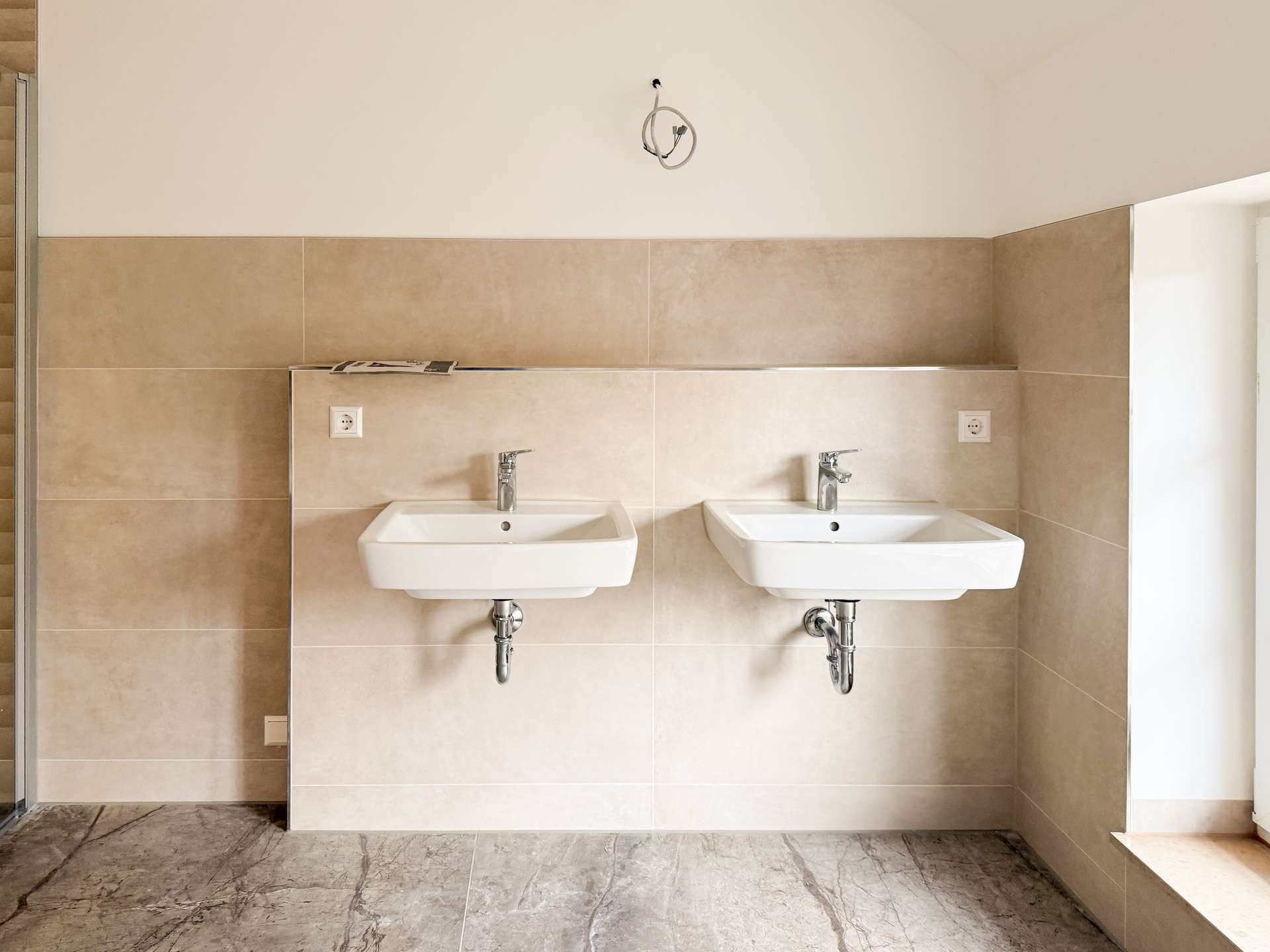 Two white wall-mounted sinks with chrome taps set in a beige-tiled wall area of a bathroom, exposed plumbing underneath and marble floor tiles.