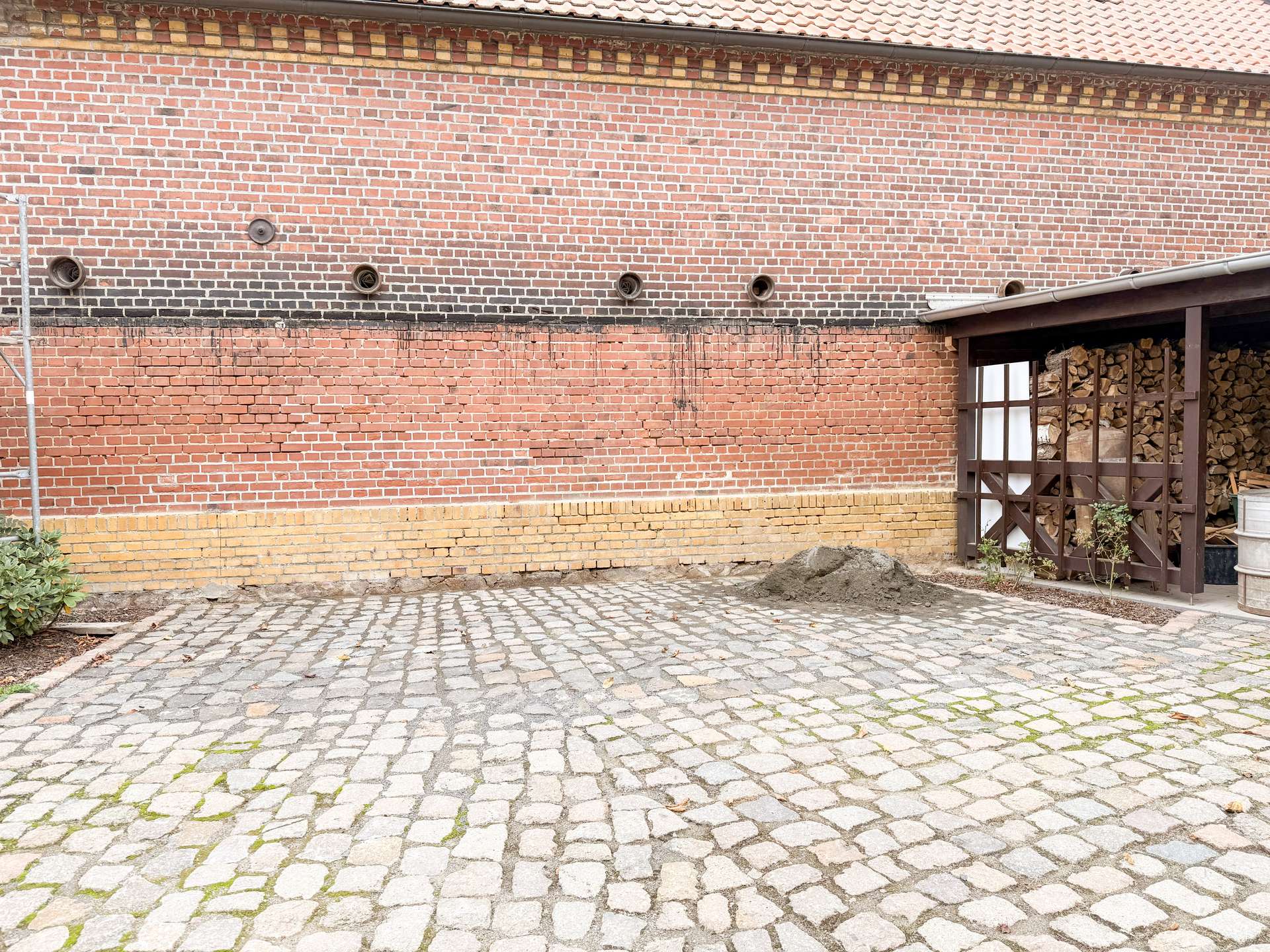 Red brick wall with four circular vents, a darker mortar band, and a woodpile storage area under a roof on the right, all on a cobblestone surface