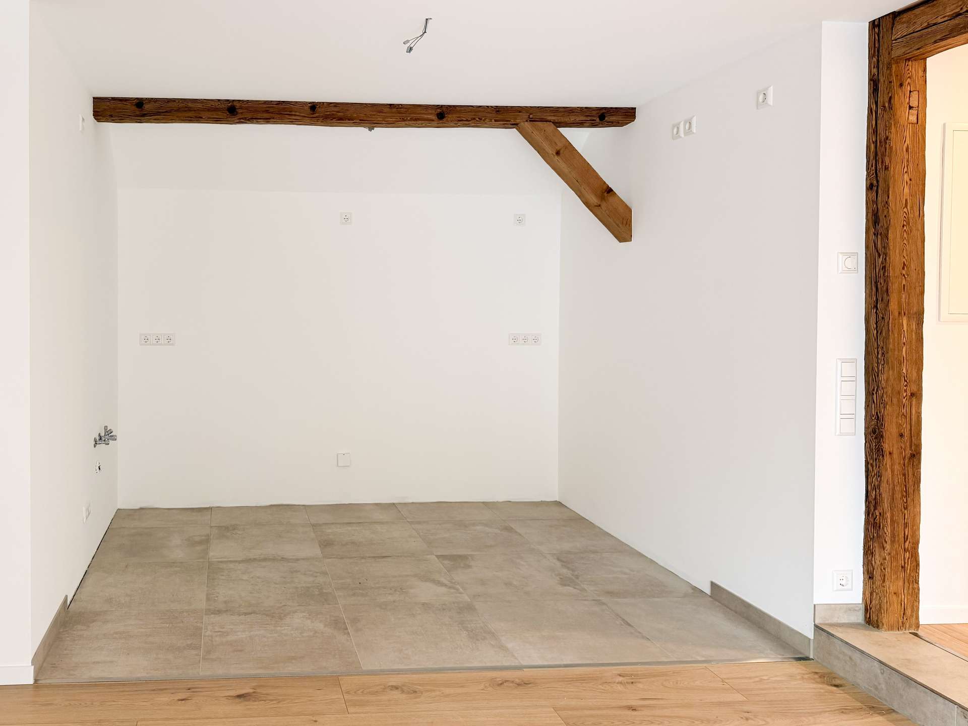 Empty white-walled room with a diagonal exposed wooden beam and tiled floor; an open wooden doorway is visible on the right side.