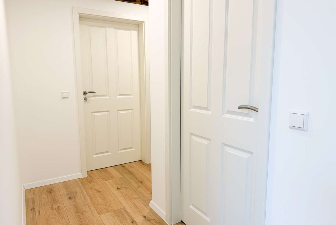 Narrow white hallway with light wood flooring, a white paneled interior door on the left and white sliding closet doors on the right Area looks clean and bright.
