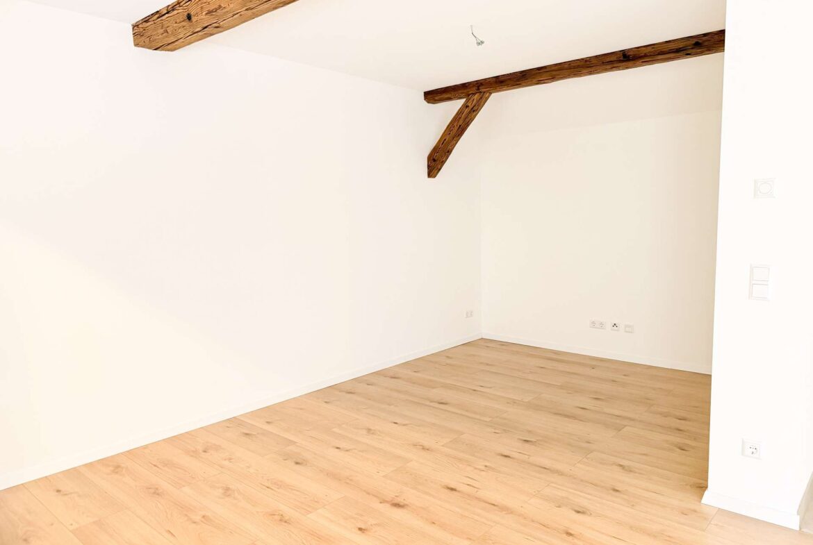 Empty bright room with white walls, exposed wooden ceiling beams, and light wood flooring.