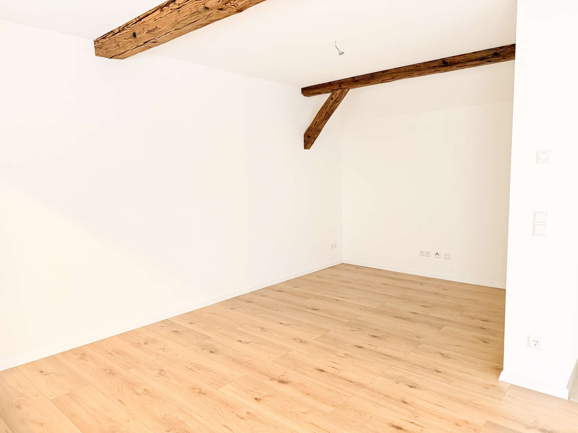 Empty bright room with white walls, exposed wooden ceiling beams, and light wood flooring.