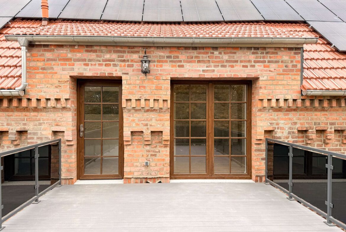 Brick house front with solar panels on a red tile roof, and two wooden-framed glass doors on a wooden deck, with a lantern between them.