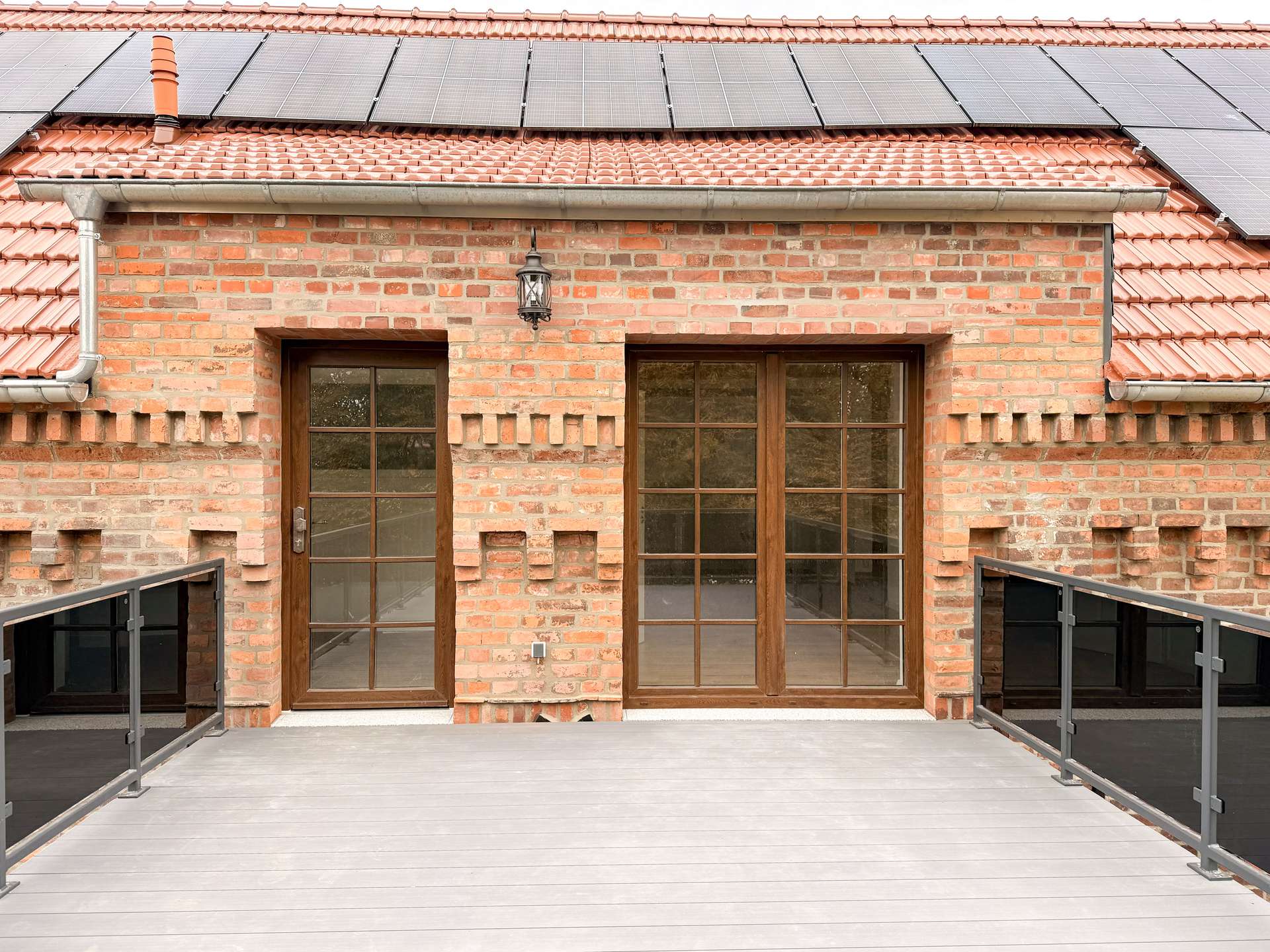 Brick house front with solar panels on a red tile roof, and two wooden-framed glass doors on a wooden deck, with a lantern between them.