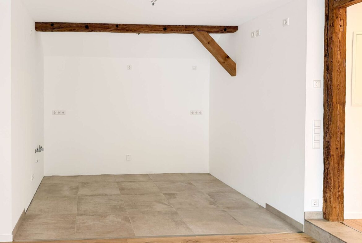 White corner of a room under renovation with two large exposed wooden beams crossing the ceiling and a diagonal brace, white walls, and a tiled floor near a wooden doorway on the right.