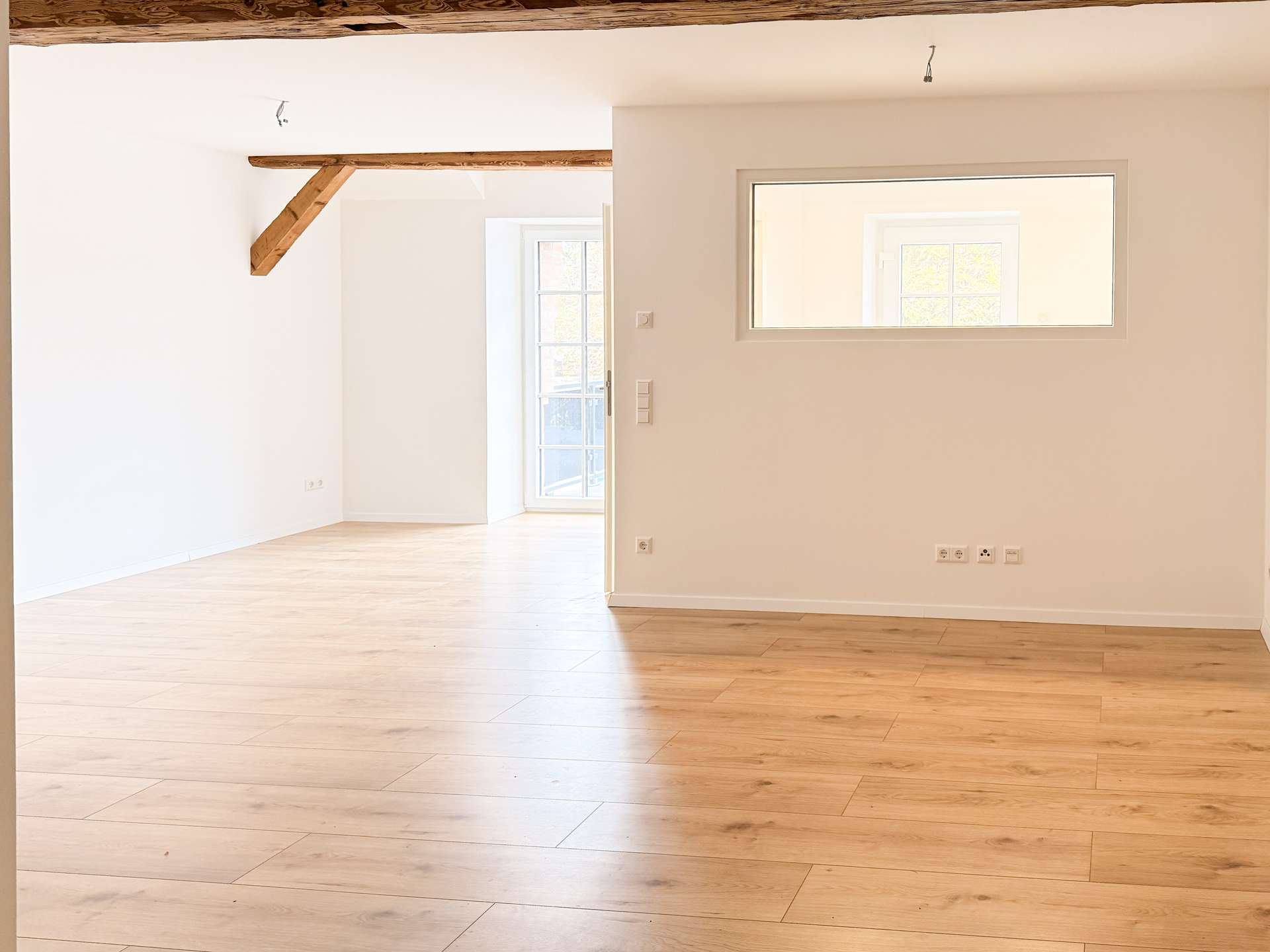 Bright empty living area with light wood flooring, white walls, and an exposed wooden ceiling beam; a doorway with glass and a rectangular window.