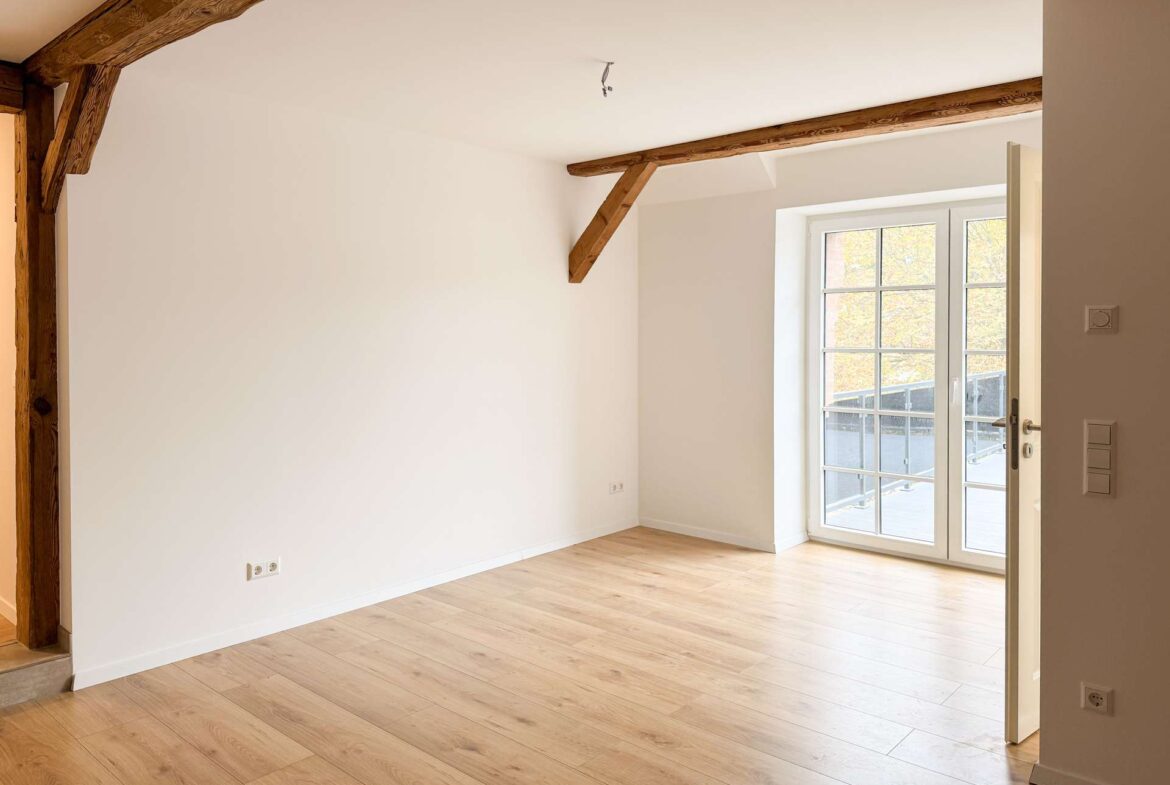 Empty living room with light wood flooring, white walls, and exposed wooden ceiling beams; glass door to balcony on the right.