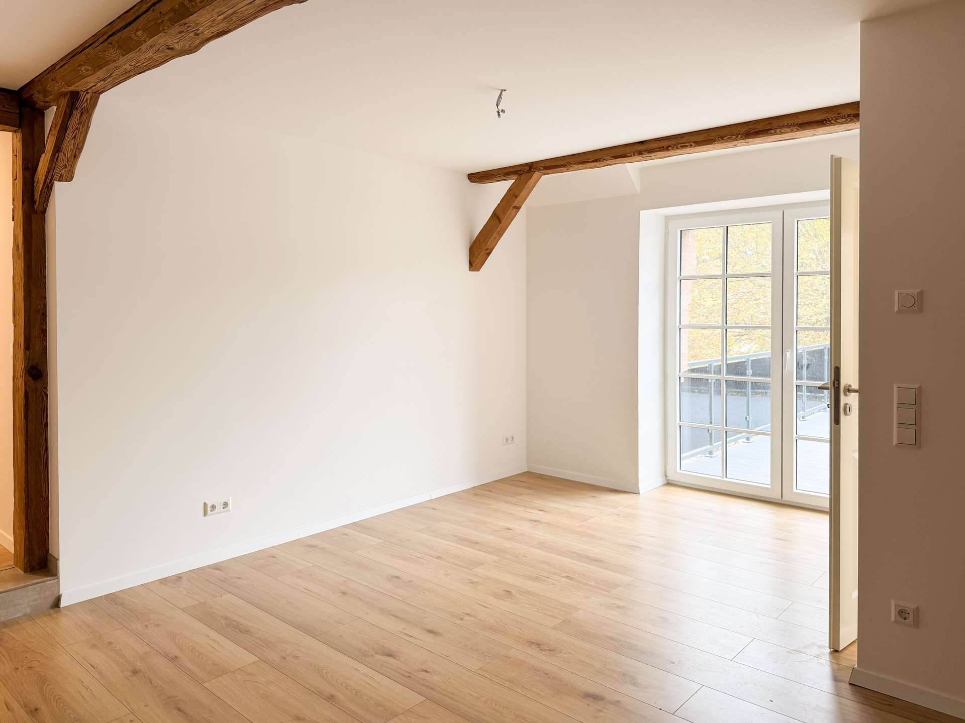 Empty living room with light wood flooring, white walls, and exposed wooden ceiling beams; glass door to balcony on the right.