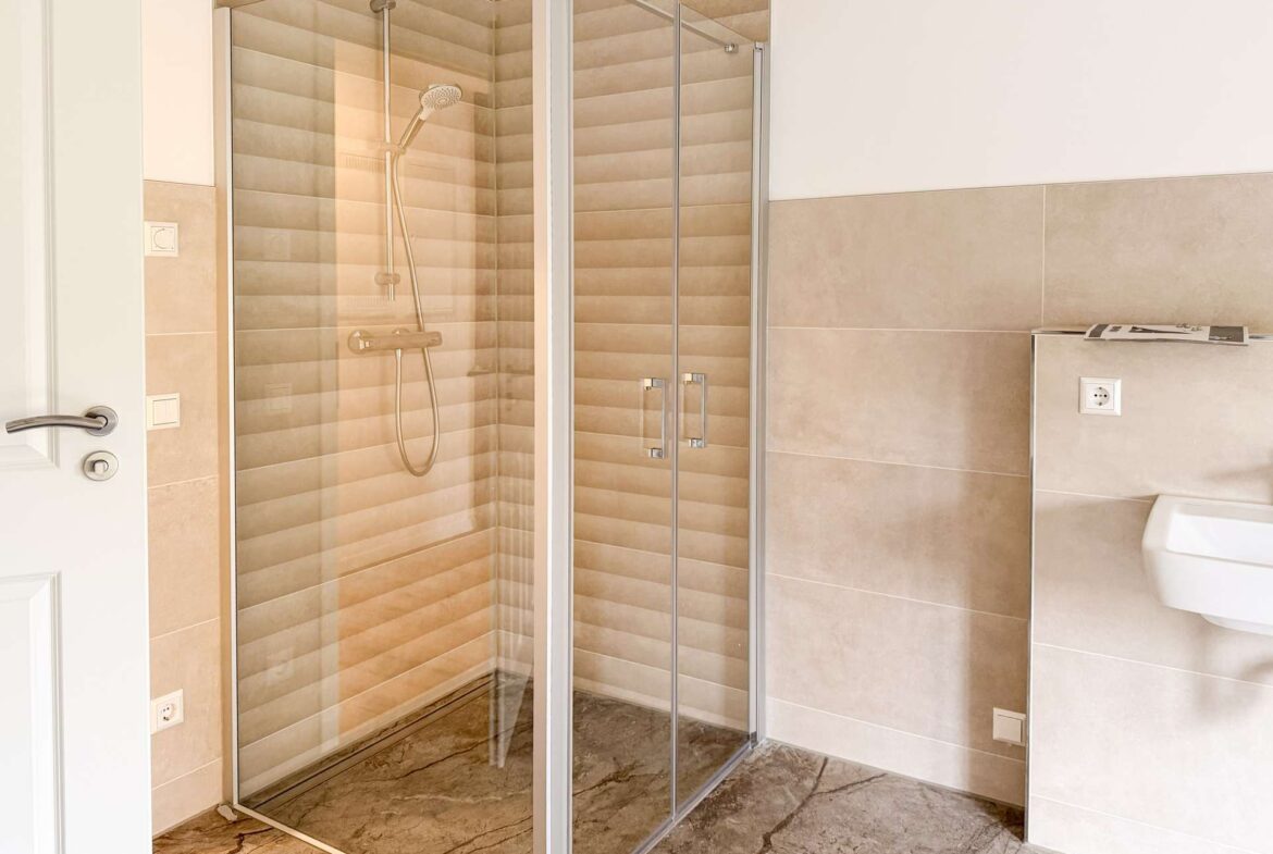 Bathroom interior with a glass-front shower, beige striped tile walls, and a marble floor; partial view of a white toilet on the right.
