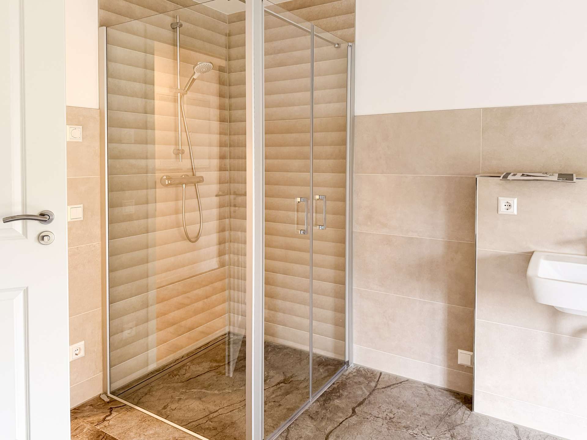 Bathroom interior with a glass-front shower, beige striped tile walls, and a marble floor; partial view of a white toilet on the right.