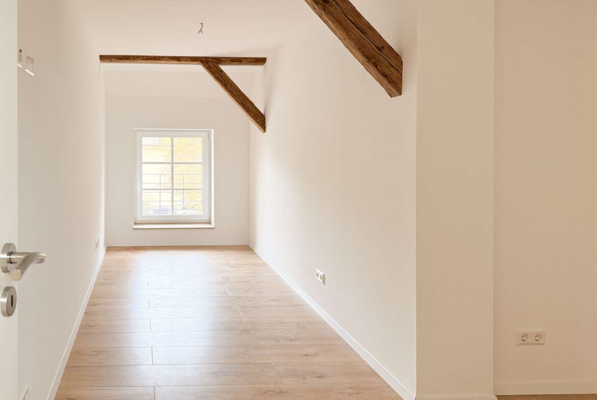 Narrow, sunlit attic room with exposed wooden beam and a small window at the far end.