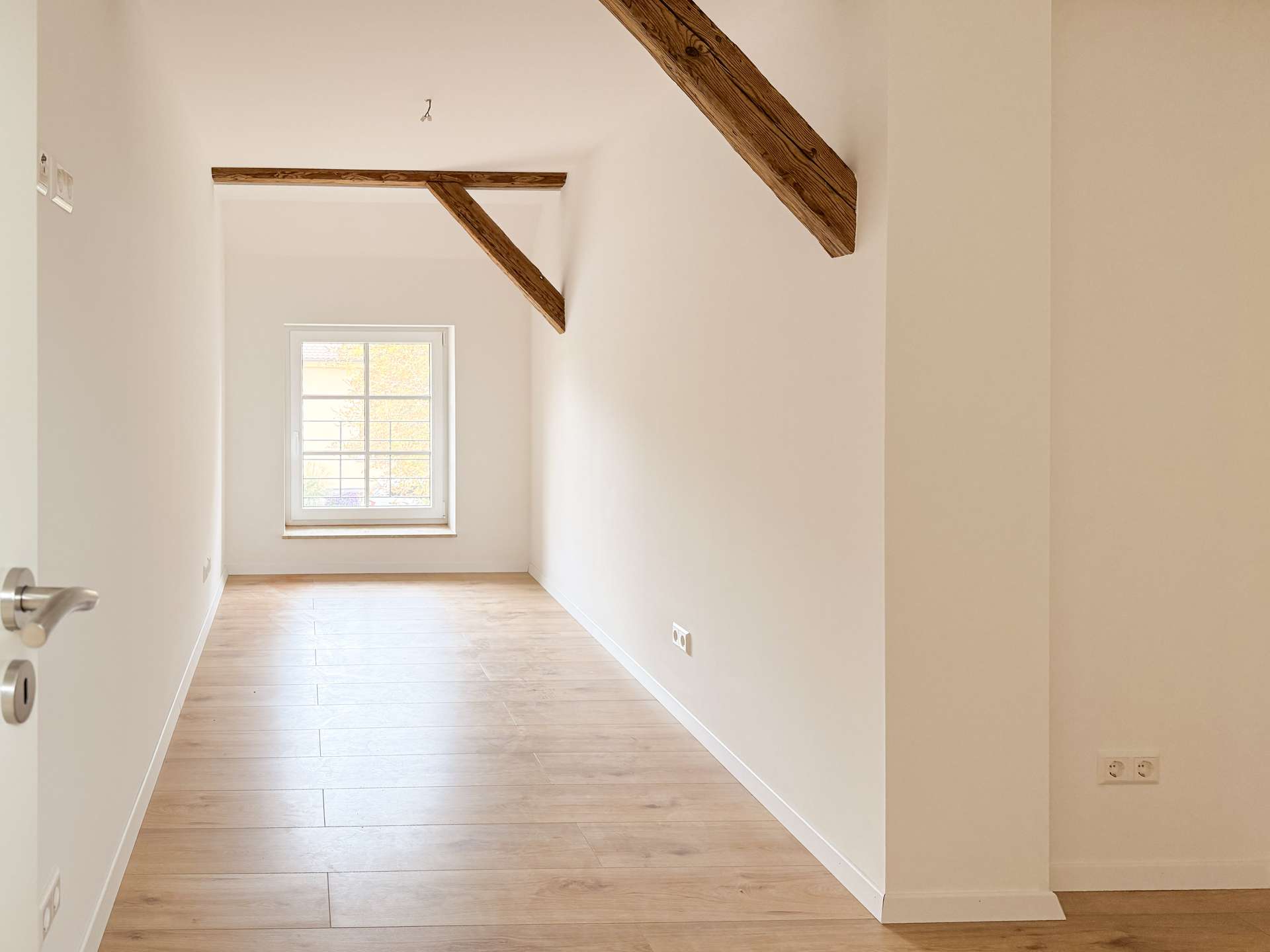 Narrow, sunlit attic room with exposed wooden beam and a small window at the far end.