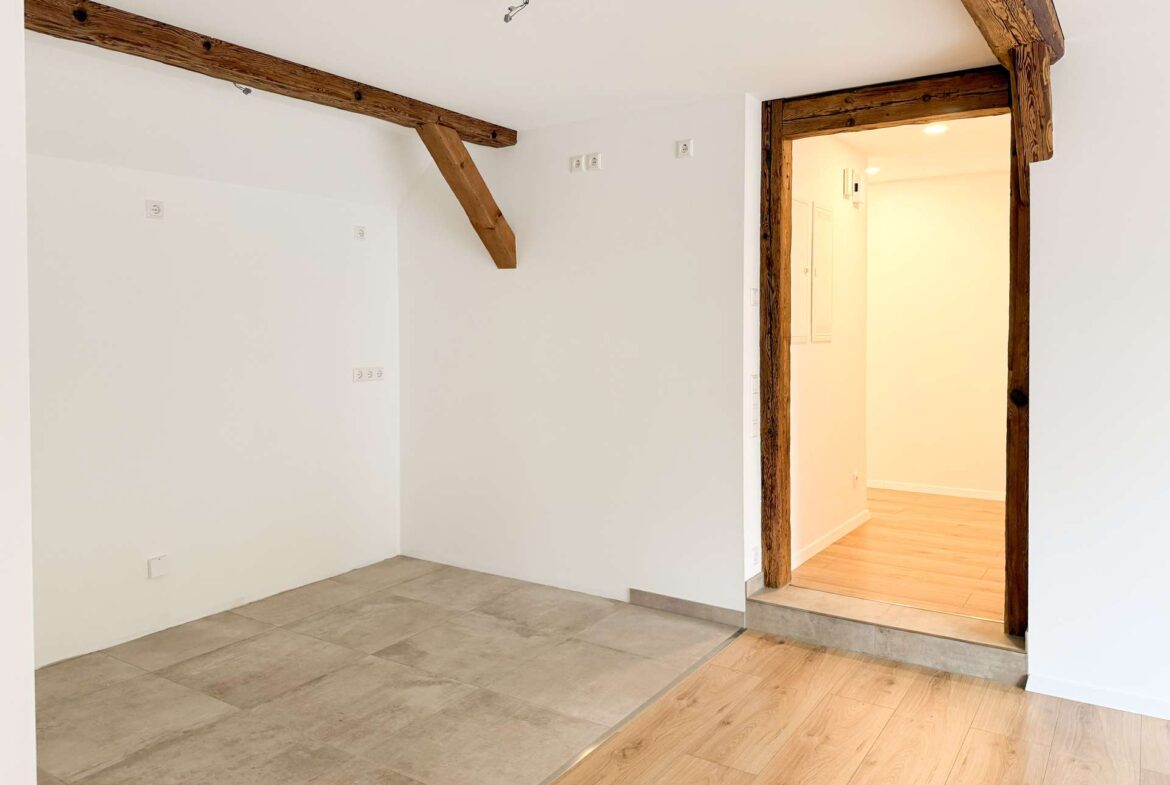 Bright interior with white walls, exposed timber beams, and a doorway framed in dark wood leading to another room.