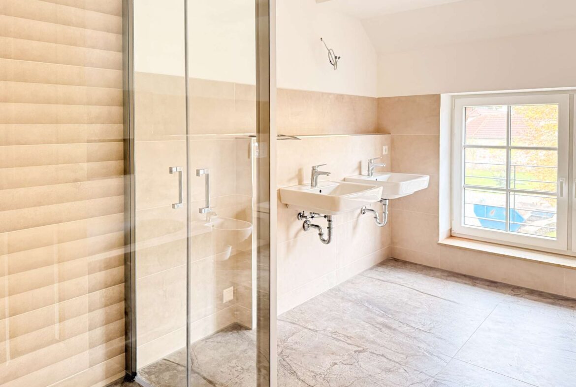 Bright bathroom with a glass-walled shower, dual white sinks, exposed chrome plumbing, beige wall tiles, and a large window showing autumn trees outside.