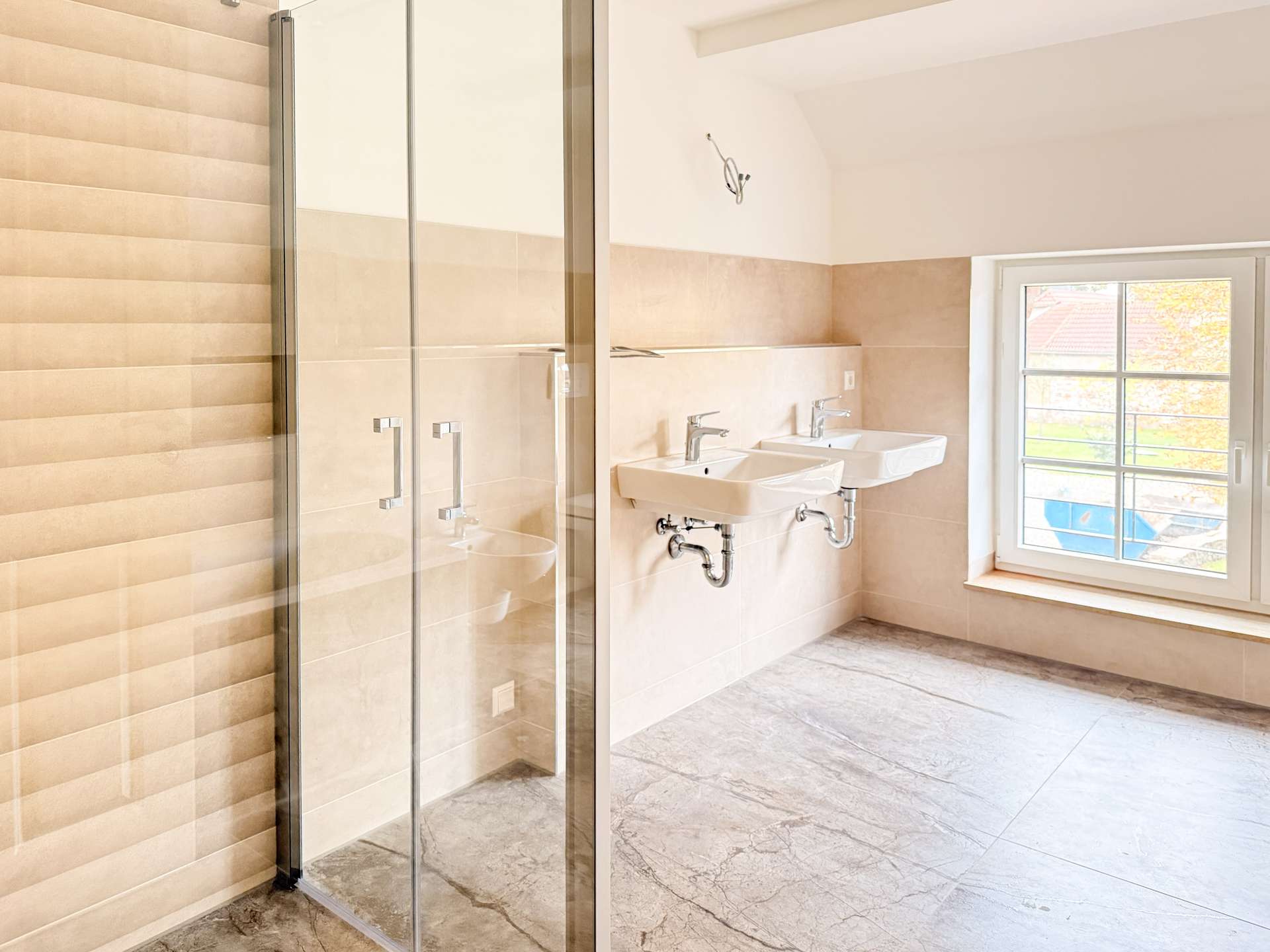 Bright bathroom with a glass-walled shower, dual white sinks, exposed chrome plumbing, beige wall tiles, and a large window showing autumn trees outside.