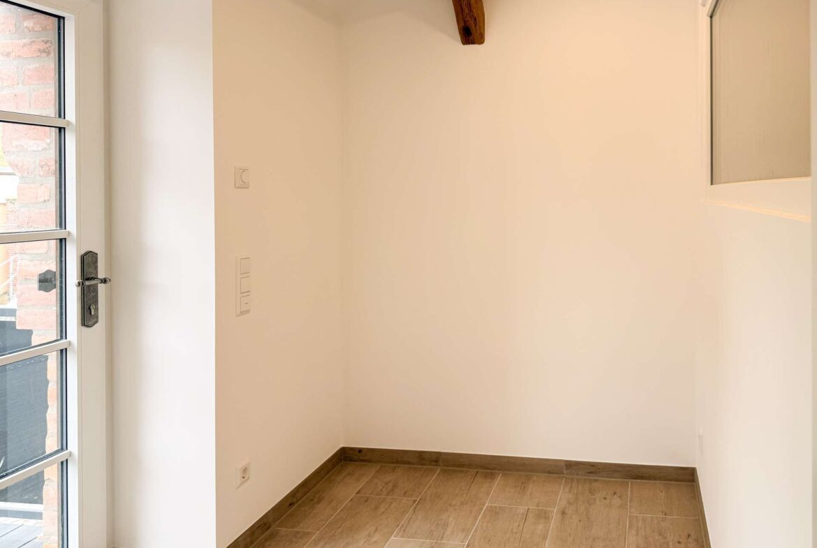 Empty entryway with a glass door on the left, visible brick exterior, white walls, a wooden ceiling beam, and wood-look tiled floor.
