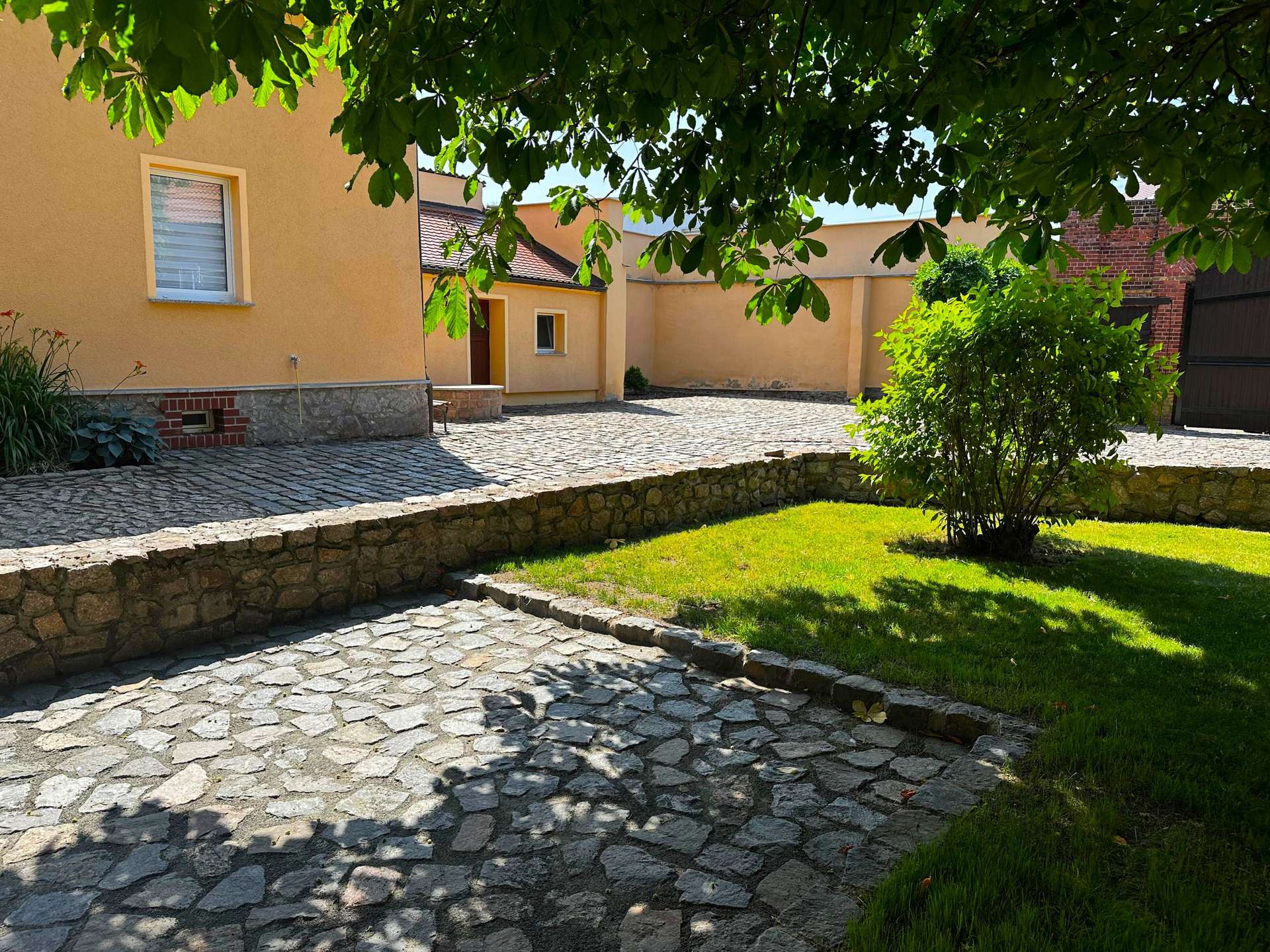 Sunny courtyard with a yellow building, cobblestone path, and green trees overhead casting shade.