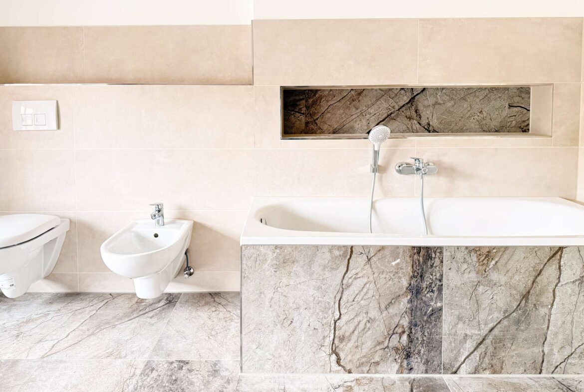 Modern bathroom featuring a white toilet and bidet on a beige tiled wall with a marble-tiled vanity and built-in shower niche edge visible above the tub area.