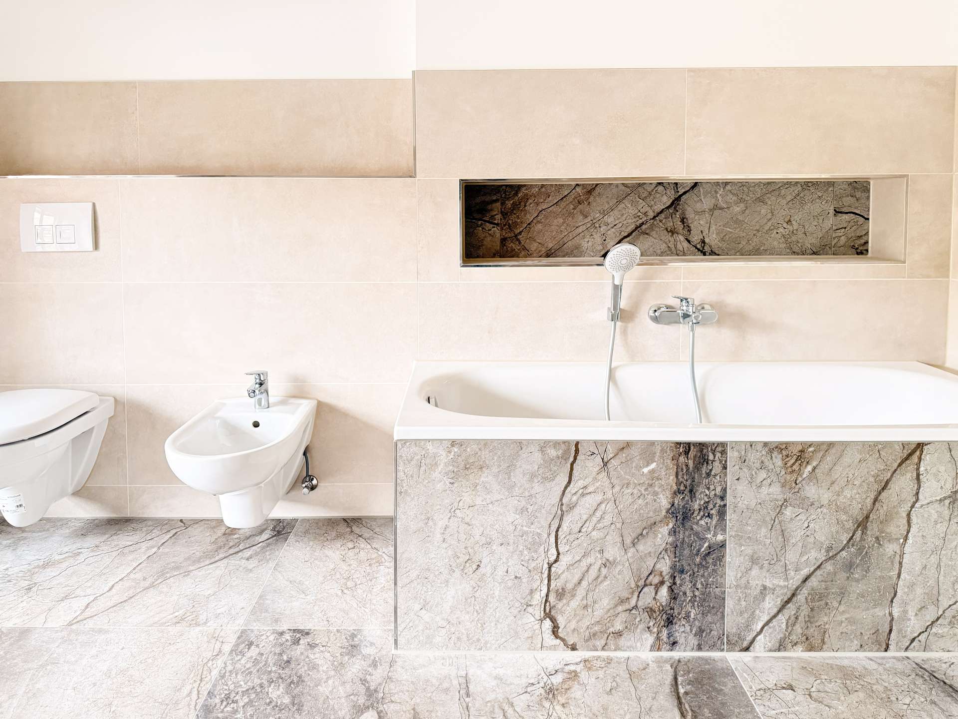 Modern bathroom featuring a white toilet and bidet on a beige tiled wall with a marble-tiled vanity and built-in shower niche edge visible above the tub area.