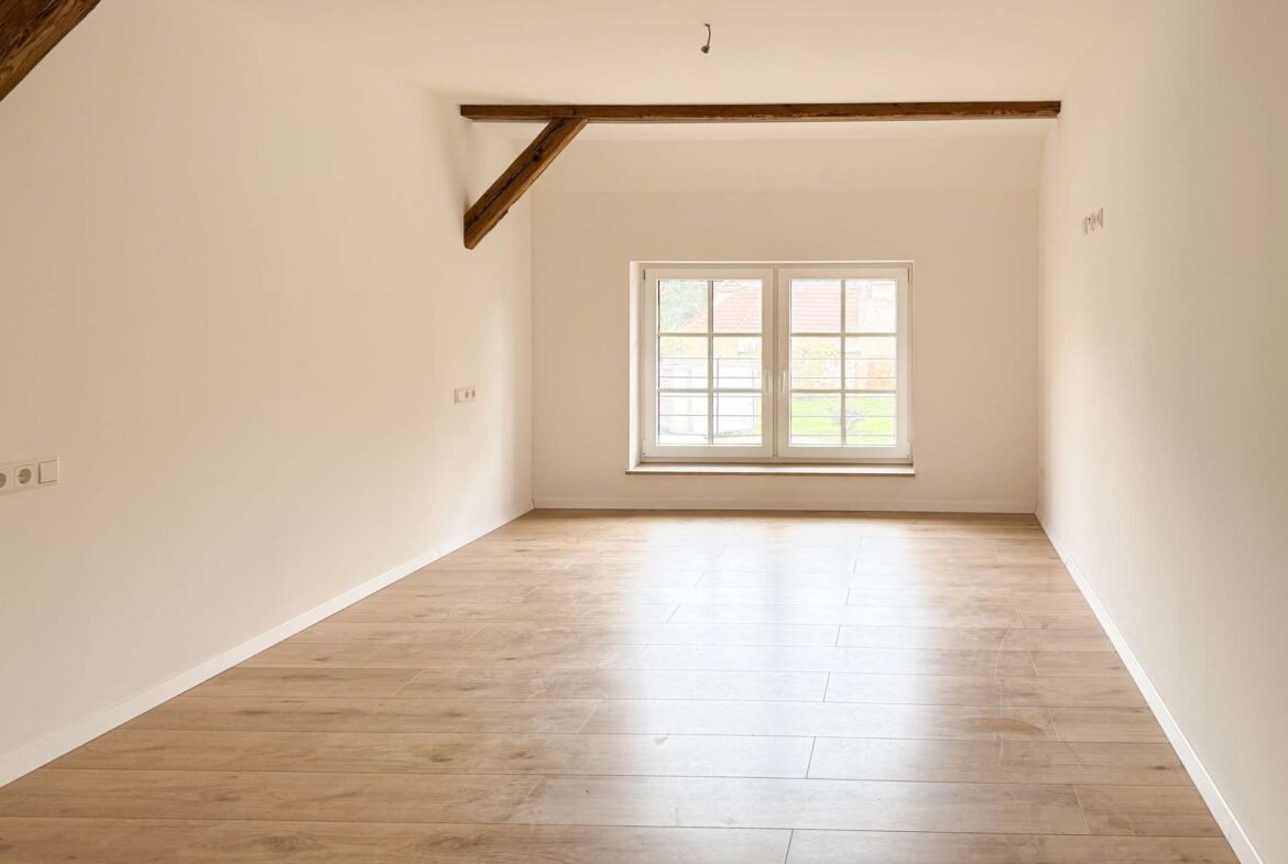 Bright empty room with light wood flooring, white walls, and a large window at the far end; a wood ceiling beam runs across the ceiling.