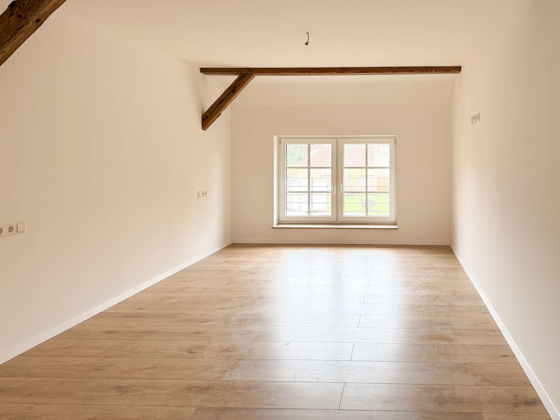 Bright empty room with light wood flooring, white walls, and a large window at the far end; a wood ceiling beam runs across the ceiling.