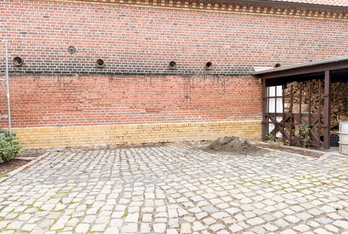 Brick wall with small circular vents above a yellow brick base; cobblestone ground in a courtyard and a woodpile shed to the right.