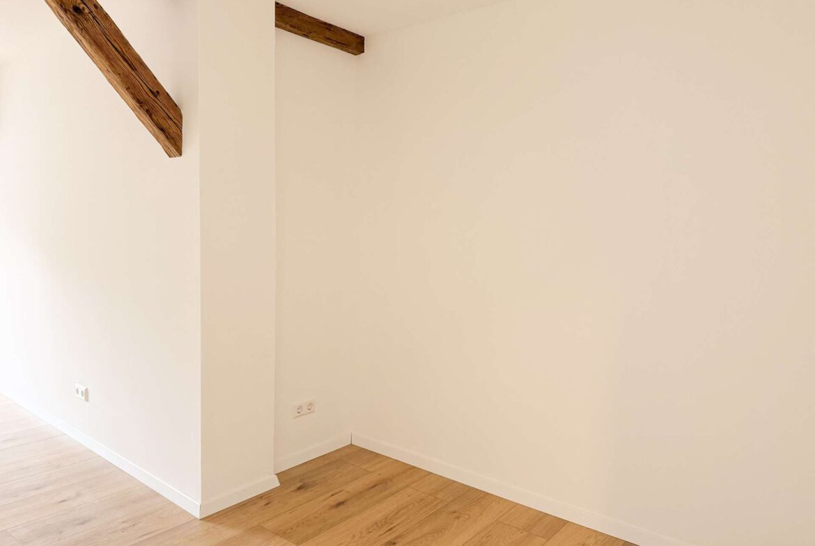Corner of a room showing exposed wooden ceiling beam, pale walls, and light wood flooring.