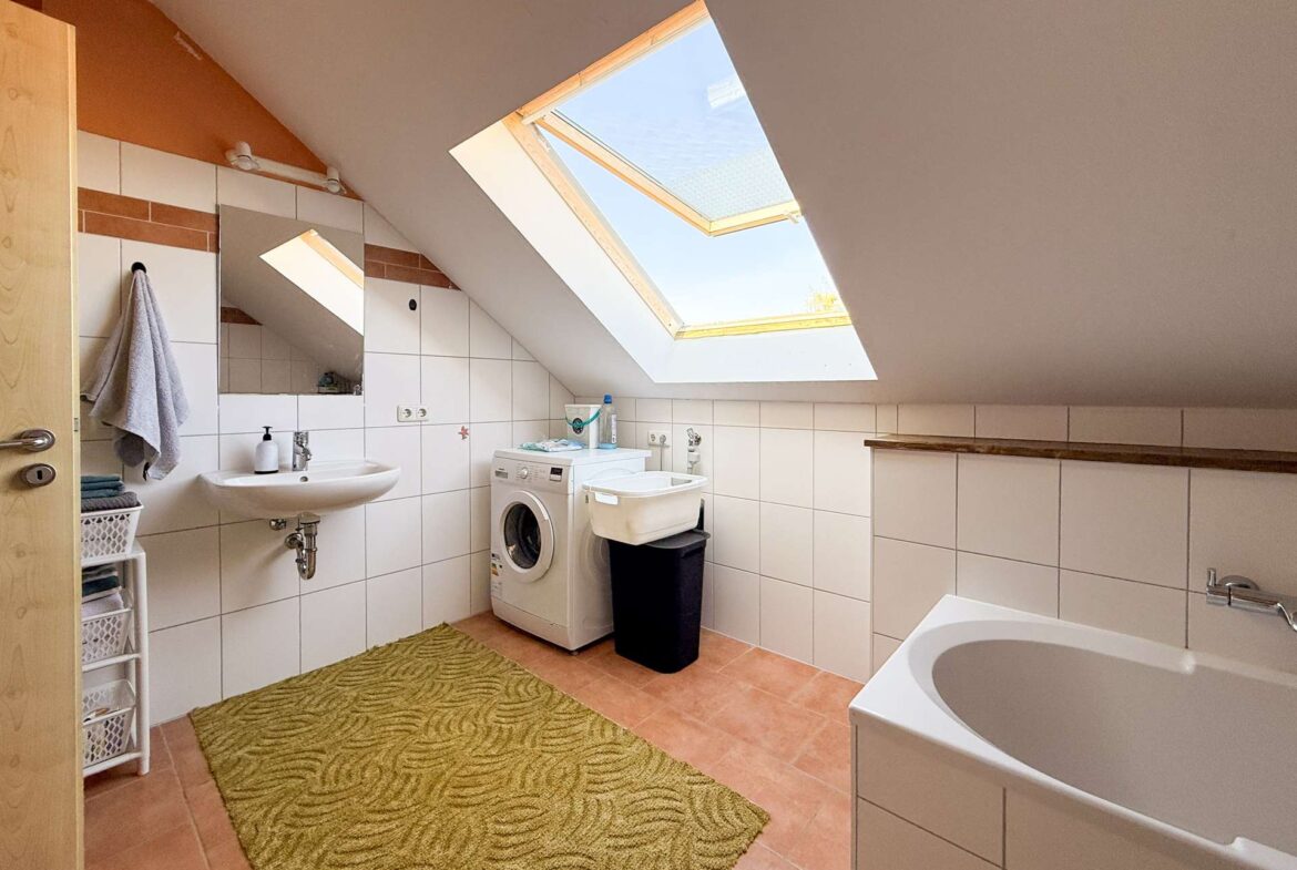Attic bathroom with a sloped ceiling and large skylight, white tiled walls, a sink, washing machine, laundry basket, and a bathtub along the right edge with a green patterned rug on terracotta floor tiles.