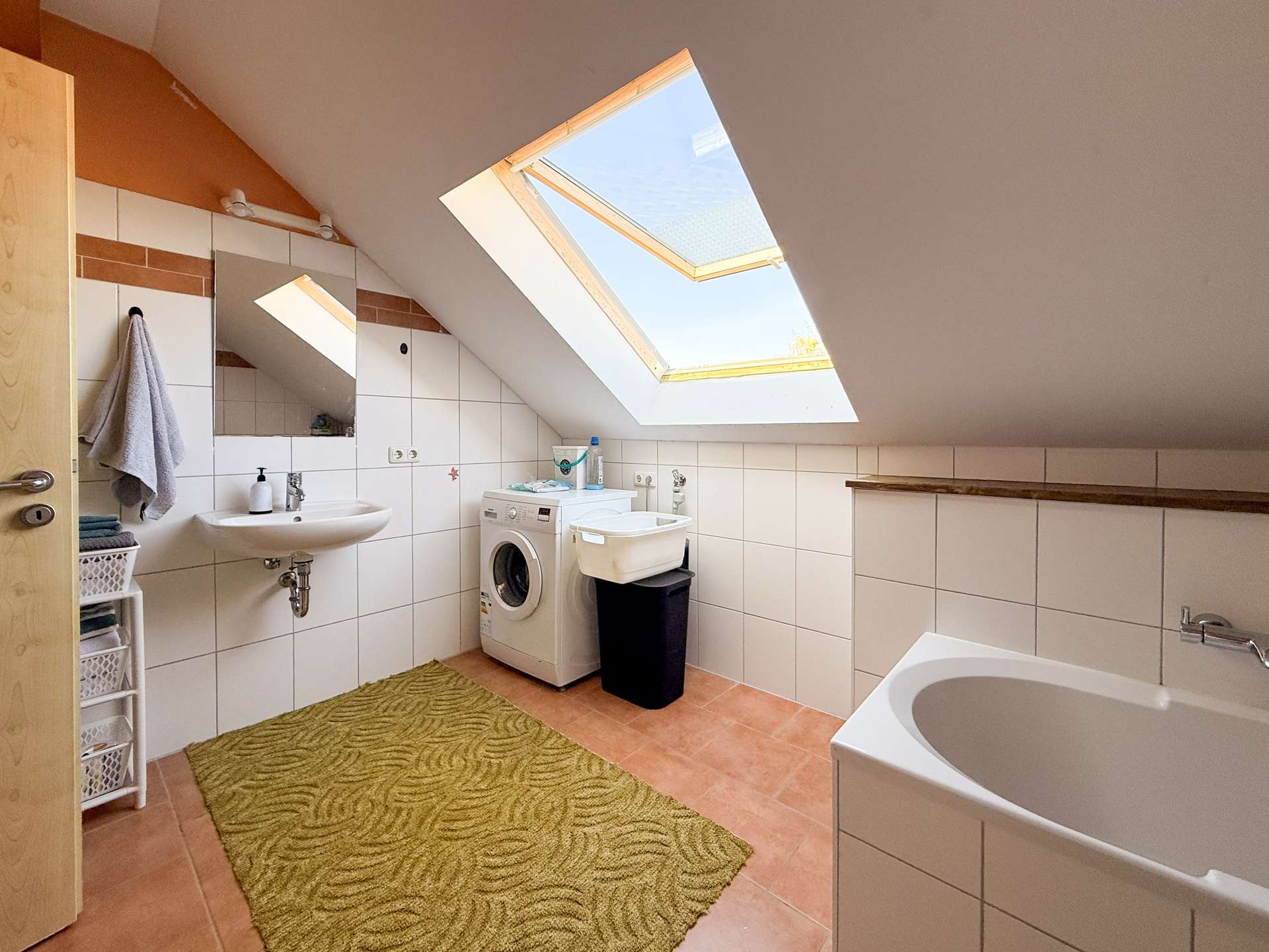 Attic bathroom with a sloped ceiling and large skylight, white tiled walls, a sink, washing machine, laundry basket, and a bathtub along the right edge with a green patterned rug on terracotta floor tiles.