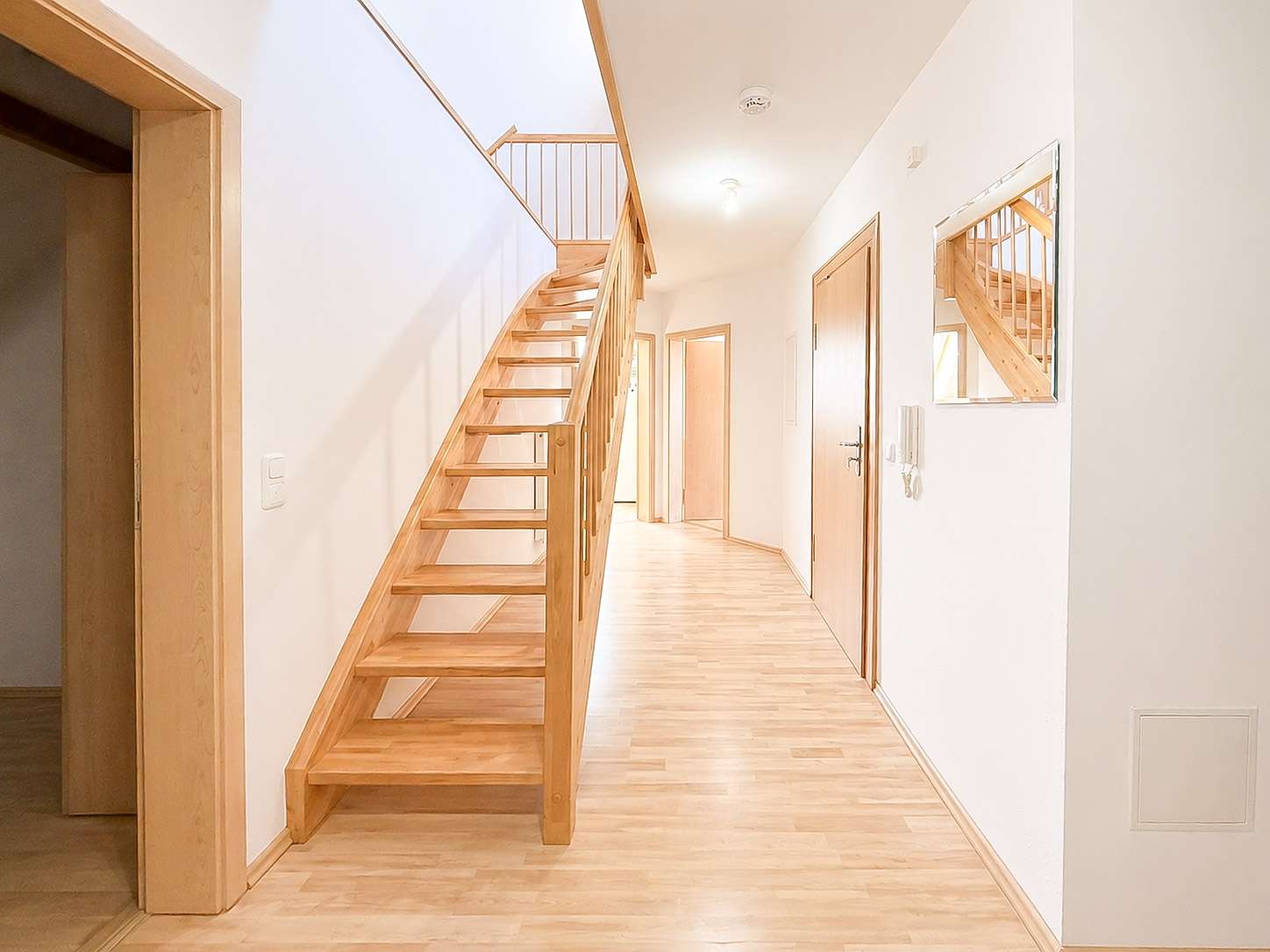 Bright modern hallway with a wooden staircase on the left, light hardwood floor, white walls, and doors along the right.