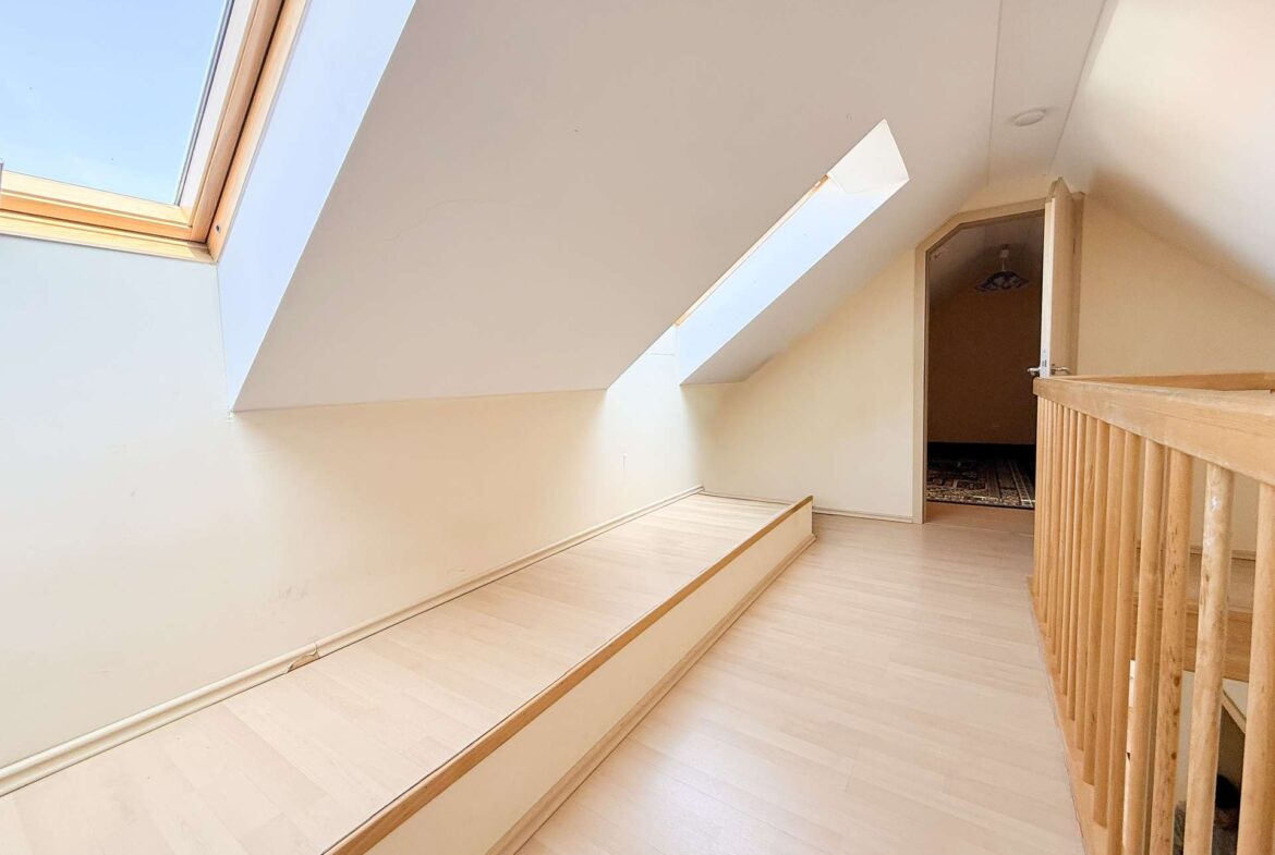 Slanted ceiling hallway with large skylights on the left, light wood flooring, and a wooden railing on the right; an open doorway at the end reveals a room with a dark patterned carpet.