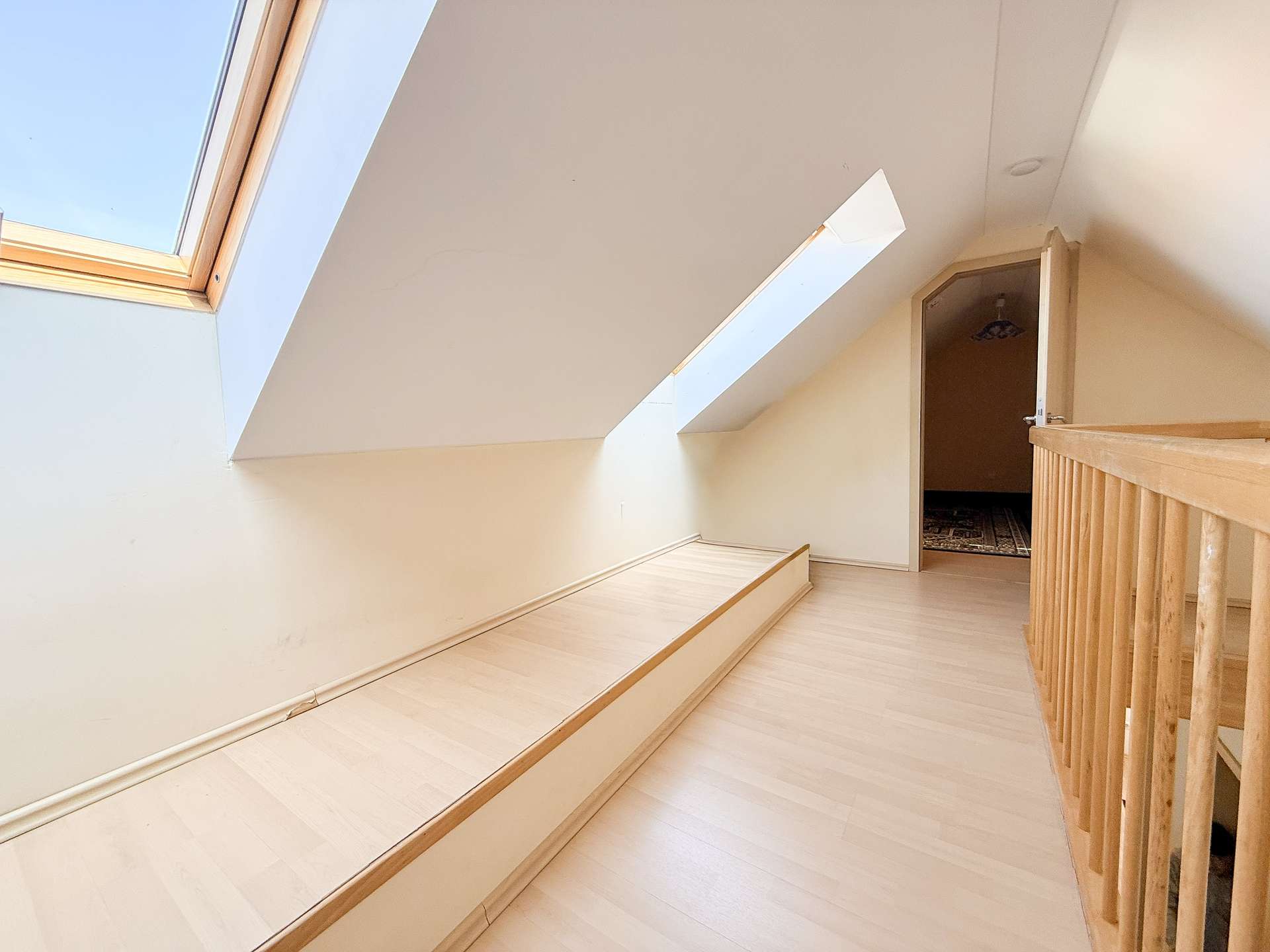 Slanted ceiling hallway with large skylights on the left, light wood flooring, and a wooden railing on the right; an open doorway at the end reveals a room with a dark patterned carpet.