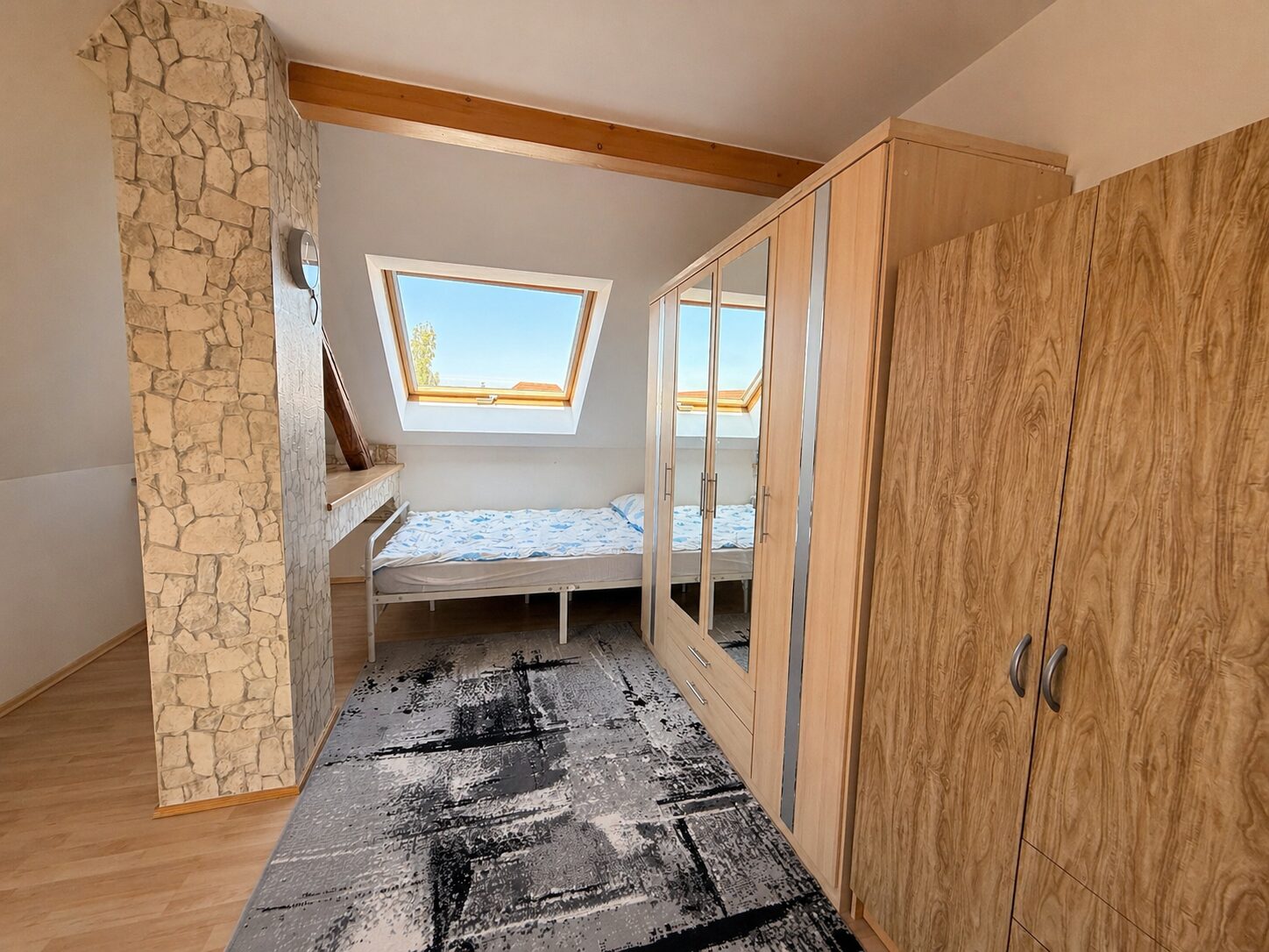 Bedroom with a single bed under a sloped ceiling and skylight window, wood wardrobe, and stone pillar feature wall.