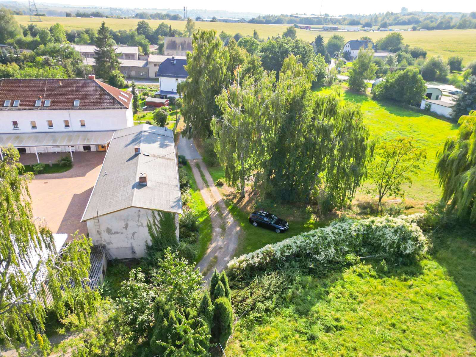 Aerial view of a rural property with white buildings, a gravel driveway, surrounding trees, and a black car parked on the grass, with fields in the distance.