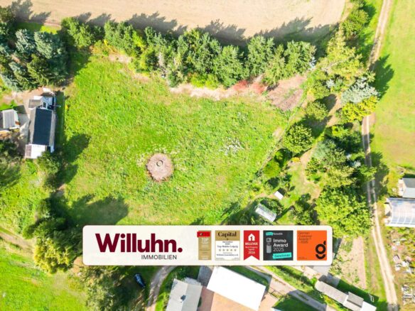 Aerial view of a grassy property bordered by trees, with a circular dirt feature in the center and buildings to the left, overlaid by a Willuhn Immobilien banner at the bottom.