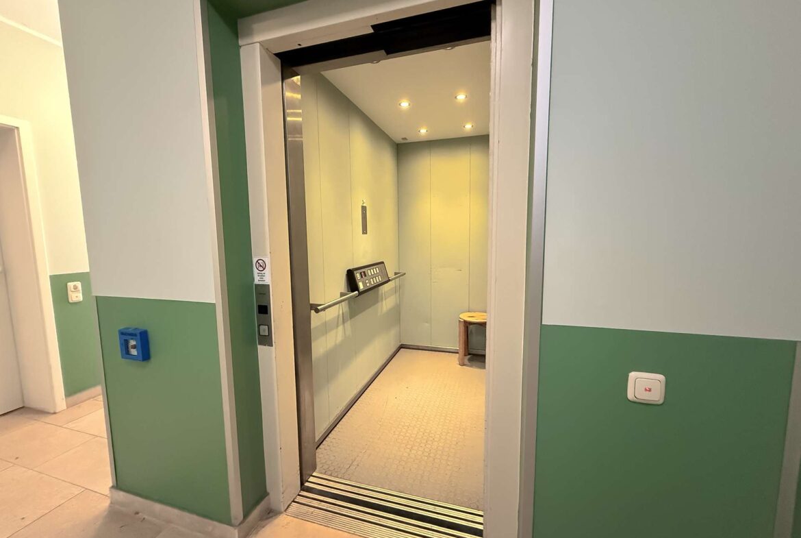 Open elevator with glass doors in a green-walled hallway, showing the control panel and a small wooden stool inside the cabin