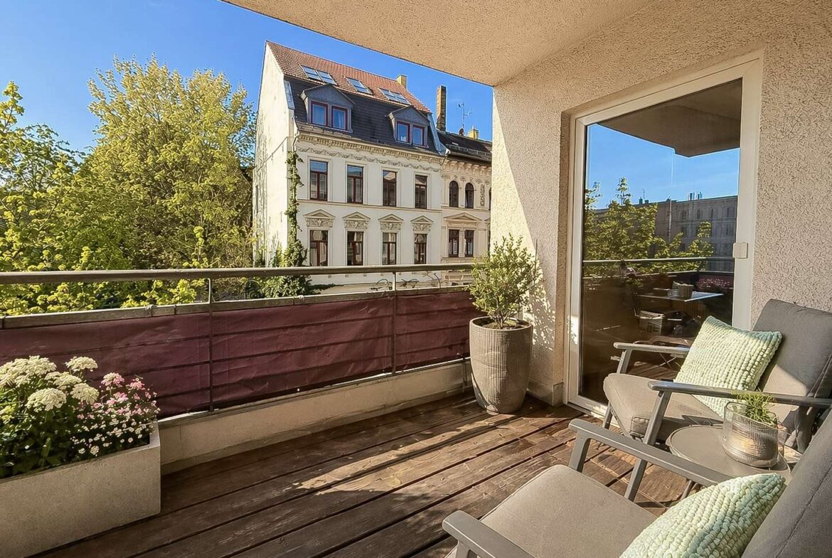 Sunny balcony with two gray chairs and mint cushions, potted plants, and a wooden deck; view of a historic white building across a railing.