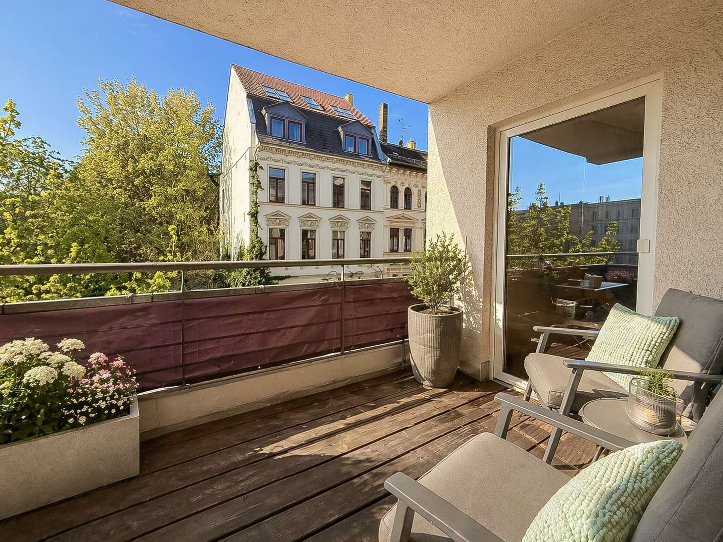Sunny balcony with two gray chairs and mint cushions, potted plants, and a wooden deck; view of a historic white building across a railing.