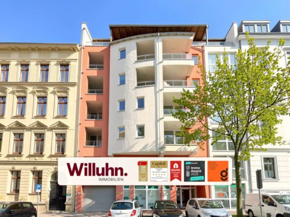 Colorful residential building fronts with a storefront sign 'Willuhn Immobilien' in the foreground and a tree on the right under a blue sky.